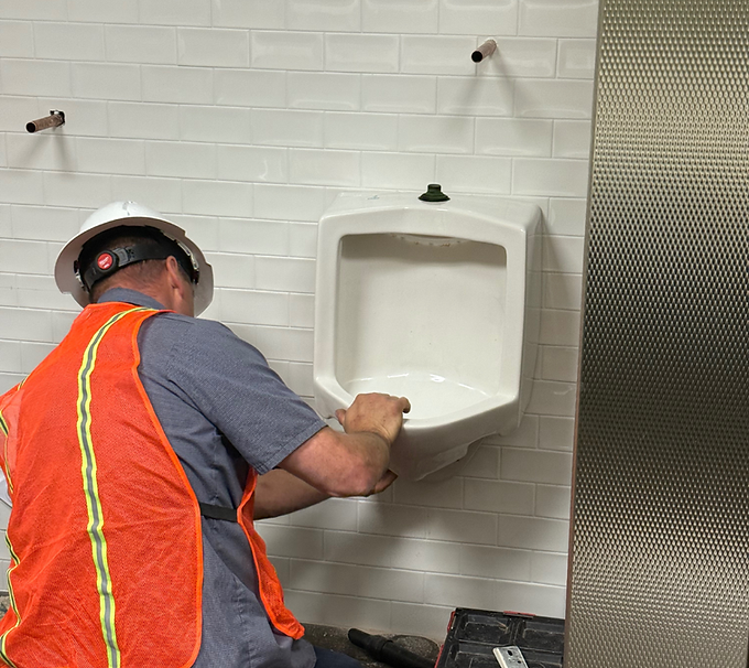 Worker in orange safety vest installing a wall-mounted urinal in a tiled restroom.