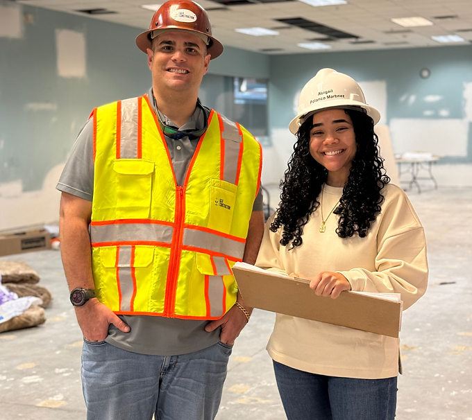 Two construction workers in hard hats and safety vests holding a clipboard in an office space