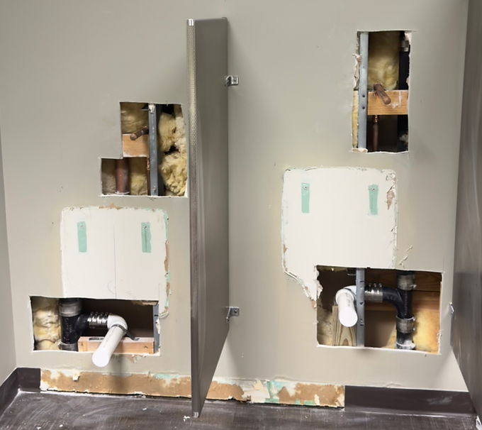 Exposed wall plumbing and open drywall cutouts around a gray utility panel in a renovation site