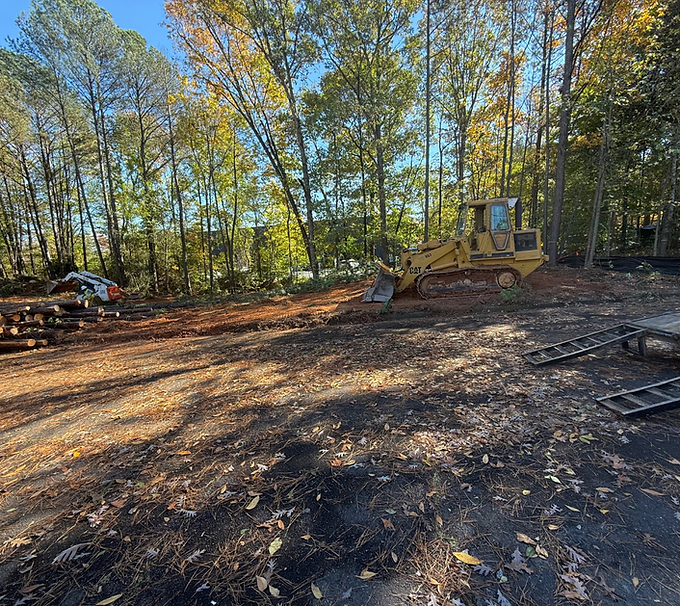 Cleared dirt lot with a yellow bulldozer beside trees in autumn sunlight