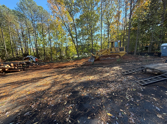 Wooded clearing with dirt ground, logs, and a yellow excavator in the distance