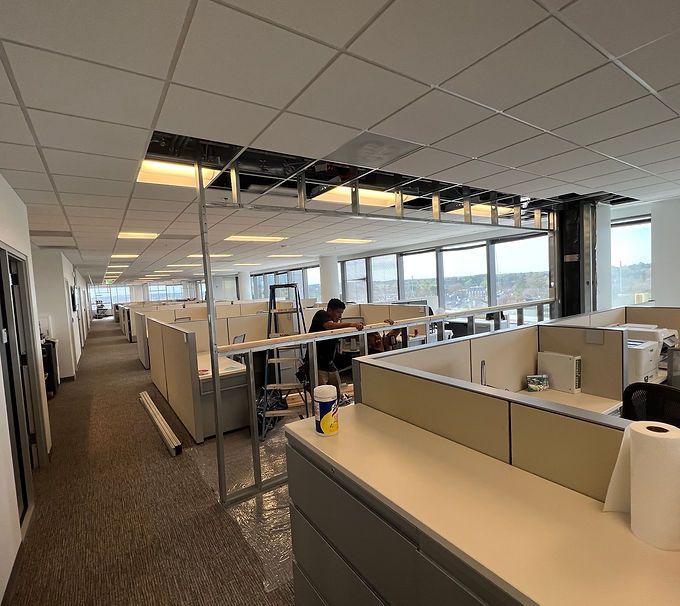 Office cubicles under a damaged ceiling with exposed wiring and a worker seated in the background