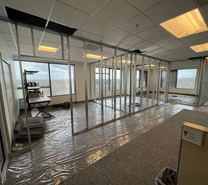 Office under construction with framed glass walls, plastic-covered floor, and ceiling lights.