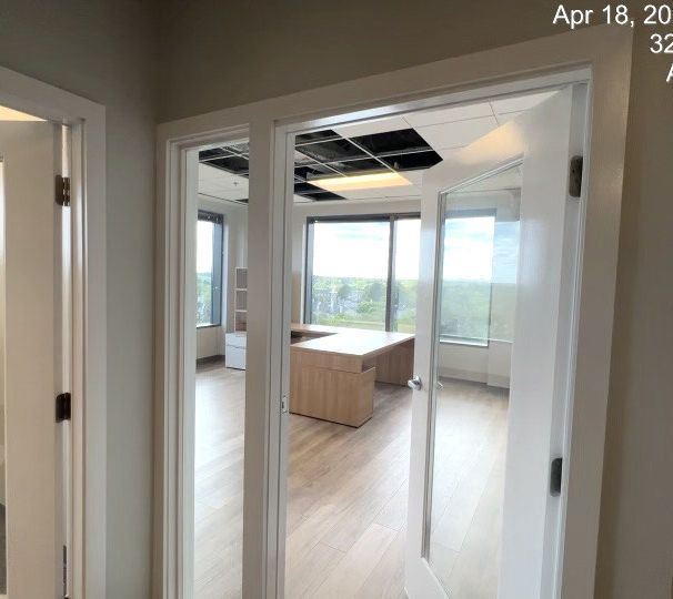 Hallway view into a bright office with glass doors, large windows, and a central wood table under ceiling panels