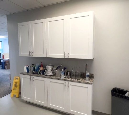 Wall-mounted white cabinets above a counter with dishes and bottles in a break room kitchen