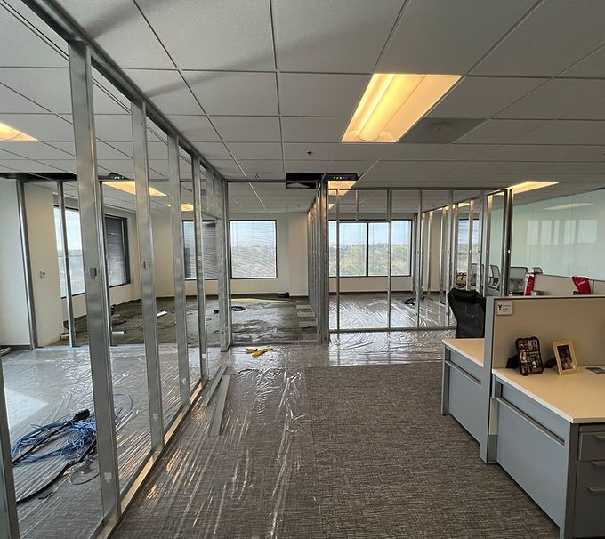 Empty office under renovation with exposed framing, carpet, and desks in a bright windowed workspace