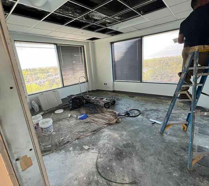 Renovation room with exposed floor, tools, and a worker on a ladder by large windows
