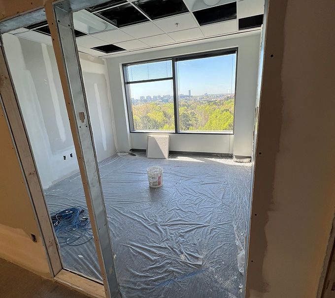 Empty room under renovation with plastic-covered floor, exposed ceiling, and large window overlooking trees and city buildings