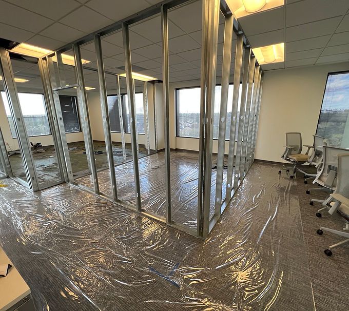 Office under renovation with exposed framing, plastic-covered floor, and chairs along the wall