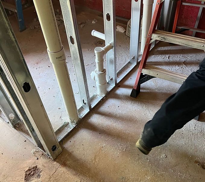 Building framing and pipes on a dusty construction floor, with a worker’s boot at right edge