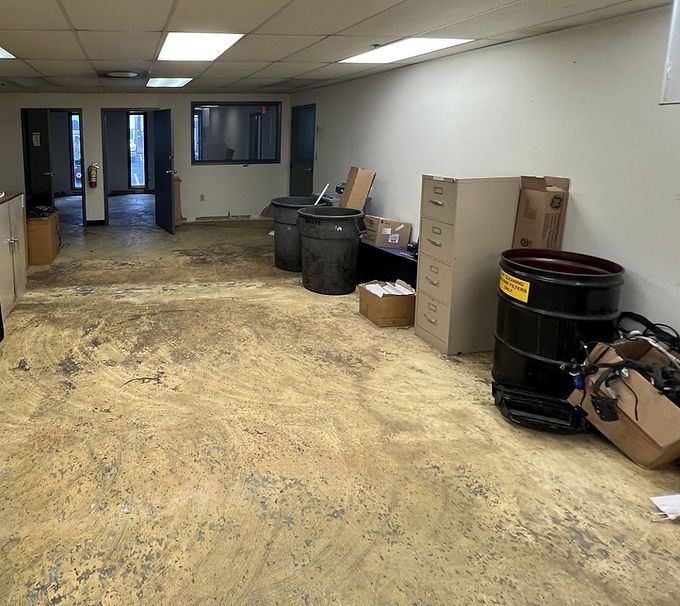 Empty office room with a damaged floor, trash bins, filing cabinets, and boxes along the wall