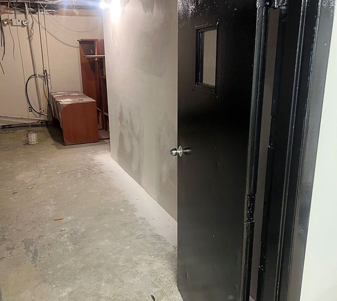 Dim unfinished hallway with a black door, concrete floor, and exposed wires near a cabinet.