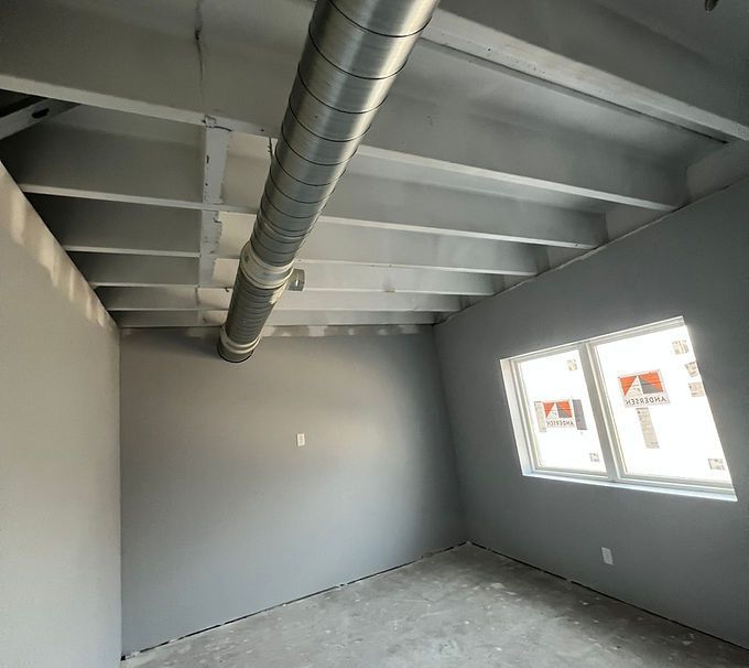 Empty gray room with exposed ductwork, white ceiling beams, and two windows letting in daylight