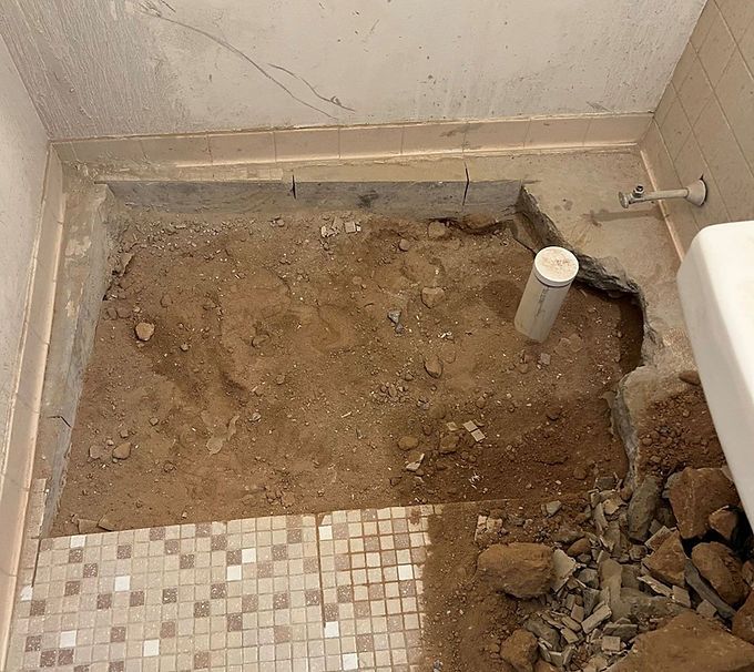 Partially demolished bathroom floor with exposed dirt, broken tile, and a white pipe near the wall