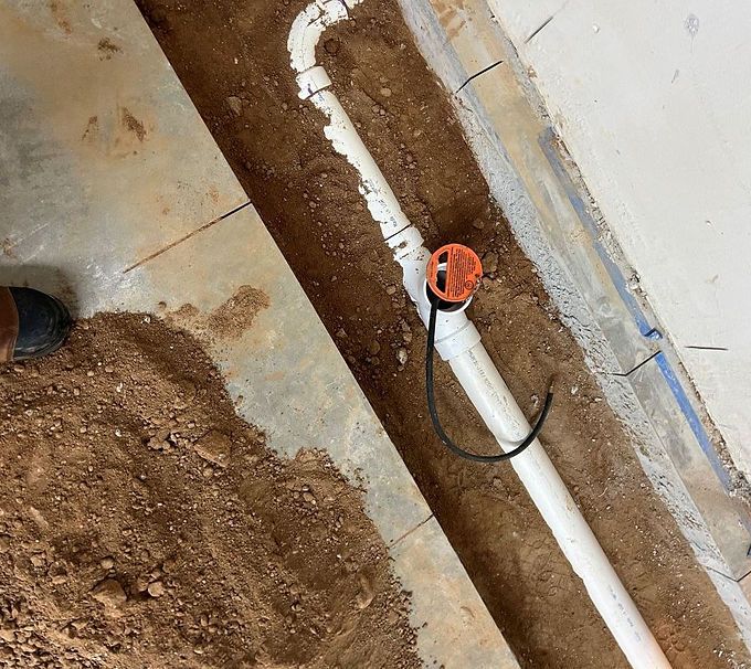 Construction trench with white PVC pipe and orange cap running along a dirt floor beside a wall.