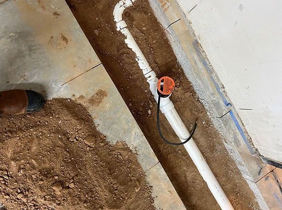 Construction trench with exposed white pipe, orange cap, and dirt along a concrete wall