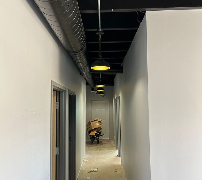 Narrow unfinished hallway with white walls, exposed ceiling ducts, and a worker near debris at the far end