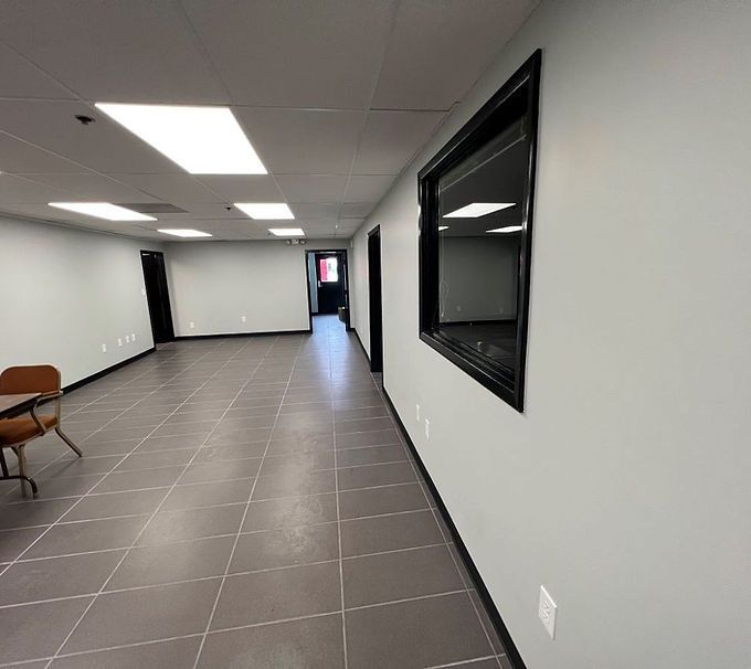 Empty white hallway with gray tile floor, ceiling lights, black trim, and a chair along the wall