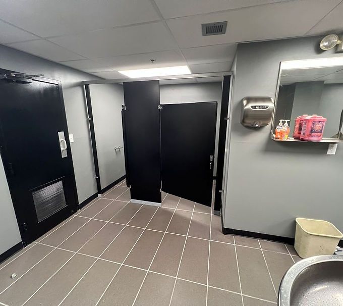 Public restroom with black stall doors, gray walls, tiled floor, and wall-mounted dispensers and trash bin.