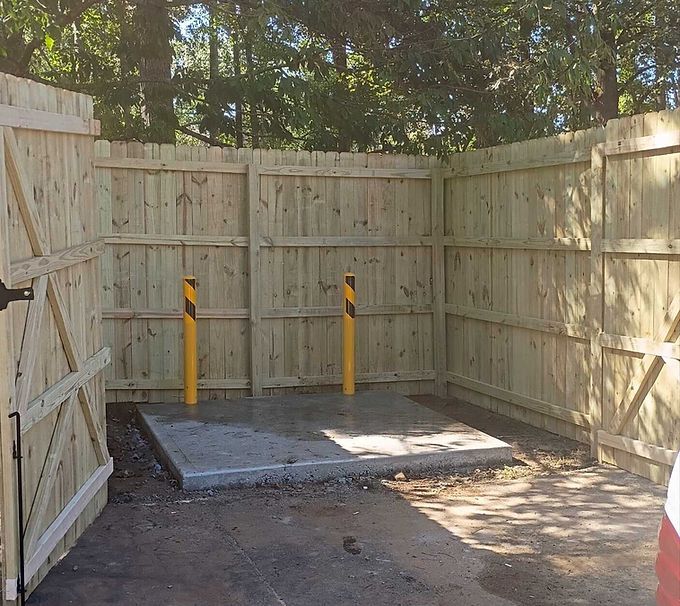 Wooden fenced enclosure with a concrete pad and two yellow-and-black bollards in a shaded yard