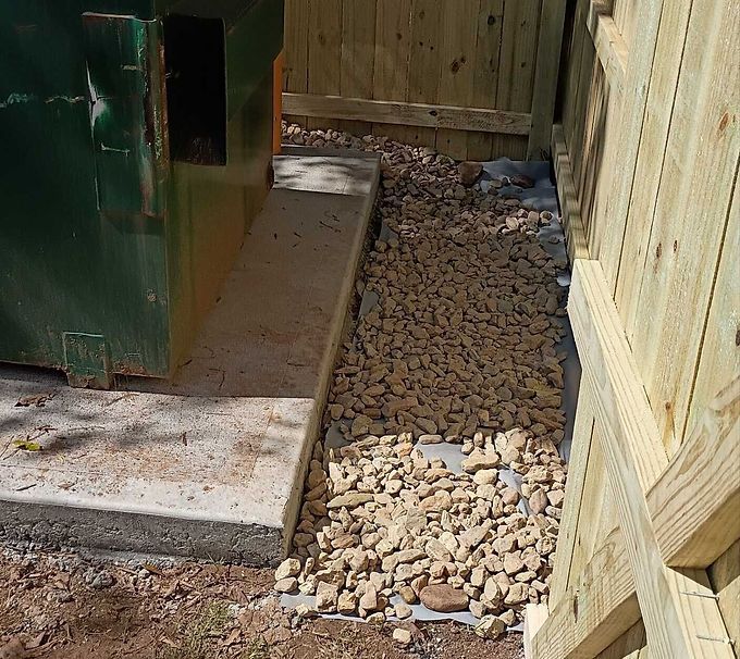 Narrow gravel path between a green structure and a wooden fence, beside concrete slabs.