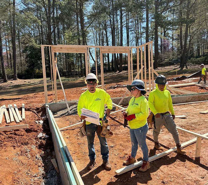 Workers in neon shirts and hard hats framing a house foundation on a sunny construction site