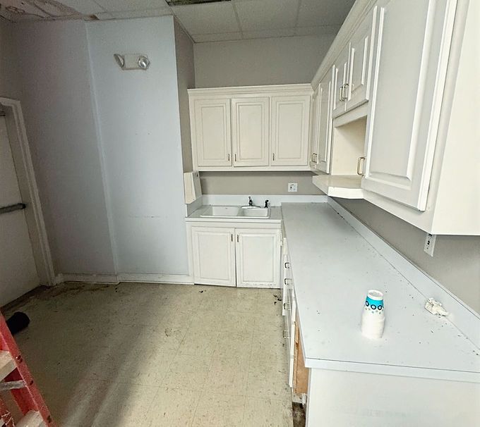 Empty white kitchen with cabinets and countertops, seen from a doorway inside a room under renovation