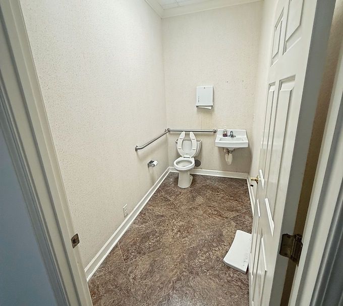 Small beige bathroom with toilet, wall grab bar, sink, and carpeted floor