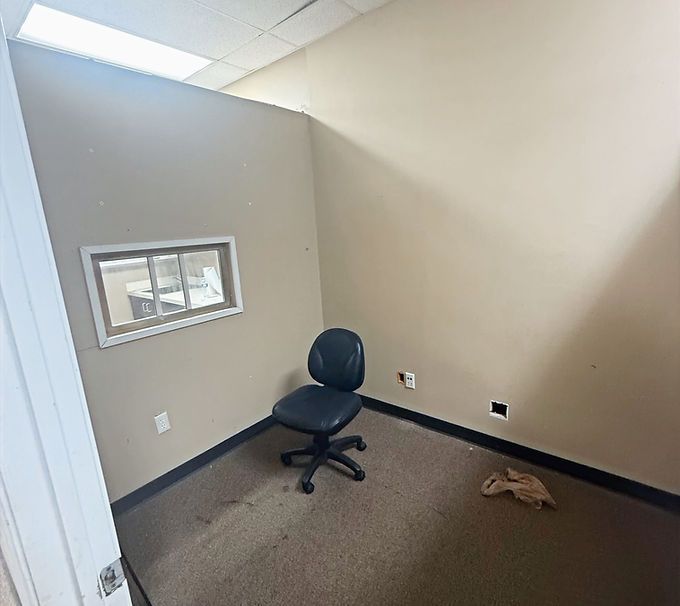 Small empty office corner with a black chair, gray walls, window, and a crumpled item on the carpet.