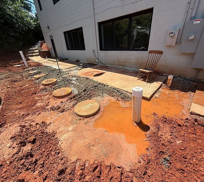 Construction site beside house with red dirt, utility pipes, and exposed round covers near windows
