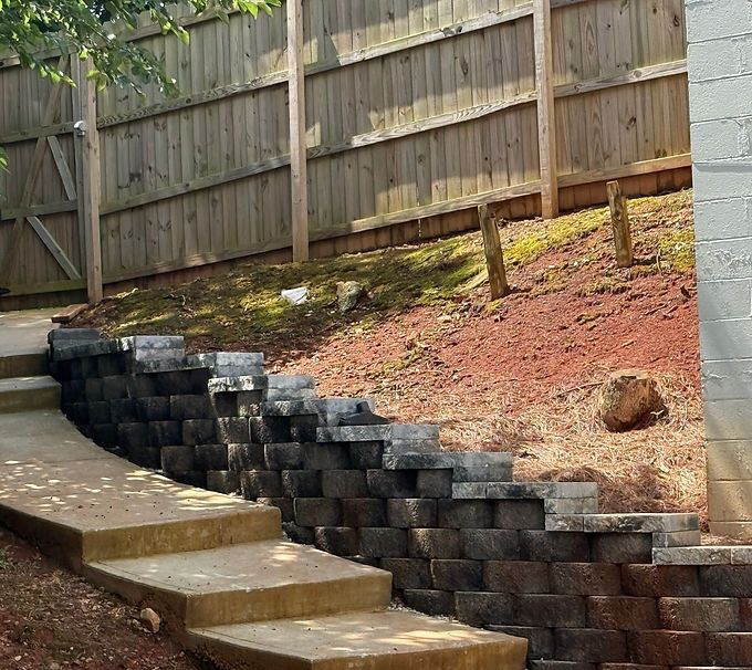 Backyard retaining wall with gray blocks beside concrete steps and a sloped red dirt hill under a wooden fence