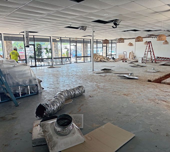 Empty office interior under renovation with exposed ceiling, tools, debris, and a ladder near the windows