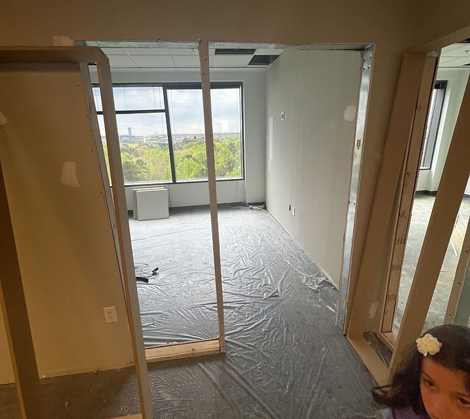 Partially renovated room with bare walls, plastic-covered floor, and large windows overlooking greenery