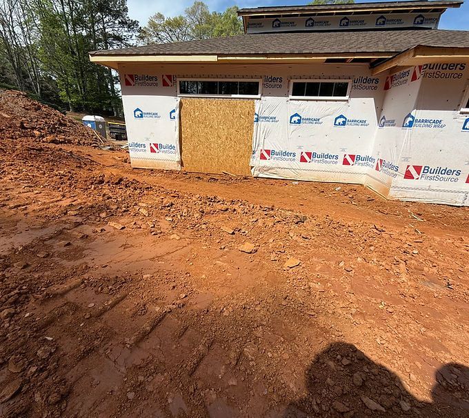 Partially built house wrapped in white Tyvek, with a boarded window and red dirt foreground