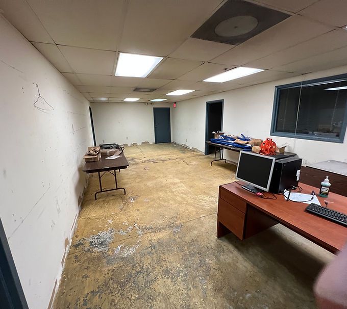 Empty office with desks, computer monitors, folding table, and stained carpet under fluorescent lights.