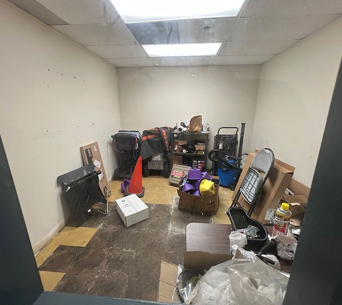 Cluttered storage room with stacked furniture, boxes, and miscellaneous items on a worn floor.