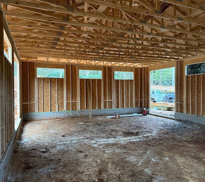 Unfinished wooden house interior with exposed framing, dirt floor, and small windows letting in daylight