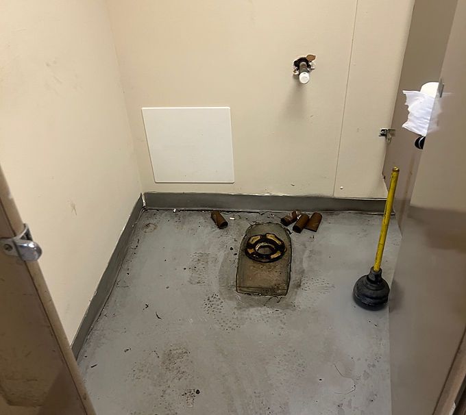 Bathroom floor under renovation with exposed toilet drain, gray subfloor, and cleaning tools.