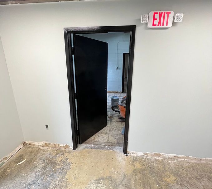 White hallway with open black door under EXIT sign, showing a dim room beyond.