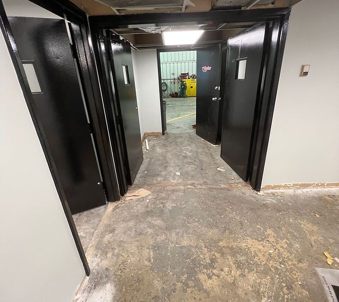Narrow industrial hallway with open black doors and a worn concrete floor leading to a bright room.