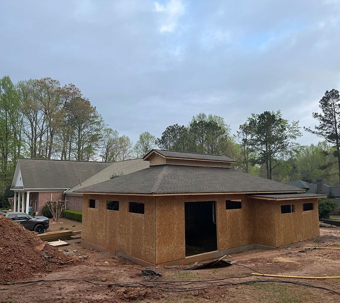 New brown building under construction on a muddy lot, with a house and trees in the background