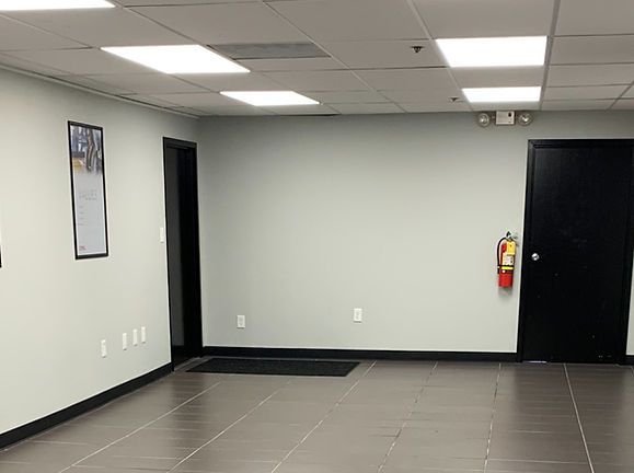 Empty office room with gray tile floor, white walls, black door, and a fire extinguisher on the wall