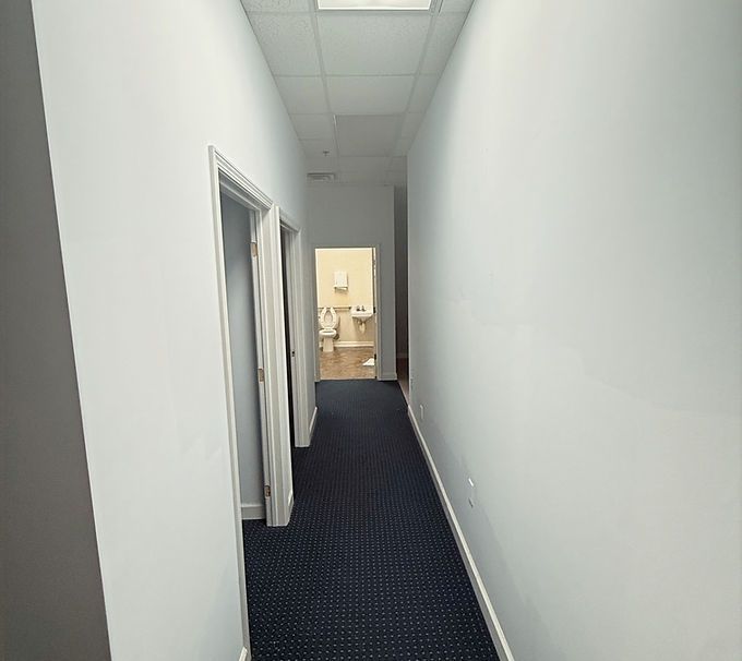 Long, narrow office hallway with white walls, dark carpet, and an open doorway at the end.