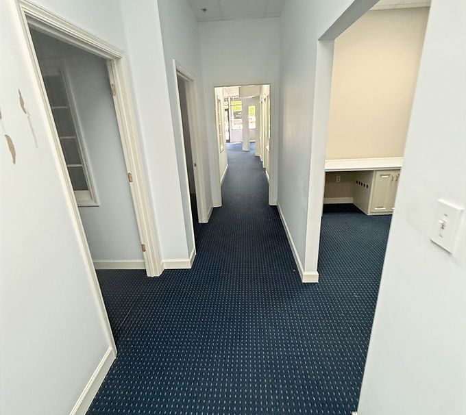 Empty office hallway with blue carpet and white walls, leading to bright rooms in the distance