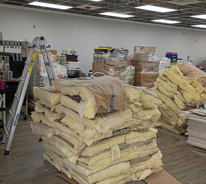 Stacks of beige insulation rolls wrapped in plastic inside a warehouse, with a ladder in the background