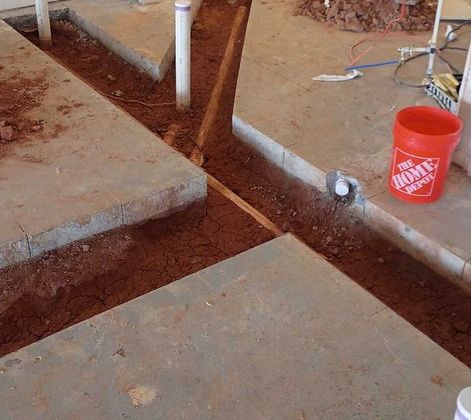 Concrete walkway with trenches and exposed white pipes at a construction site, with an orange bucket nearby