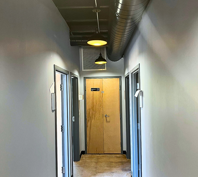 Narrow office hallway with gray walls, exposed ceiling ductwork, and a wooden door at the end