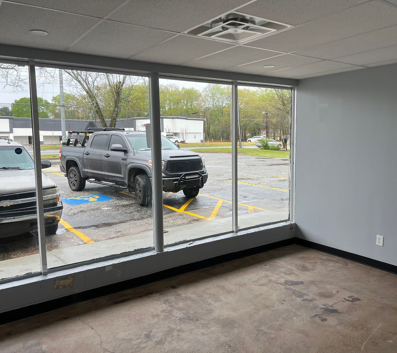 Empty storefront interior with large windows facing a parking lot and two parked trucks outside.