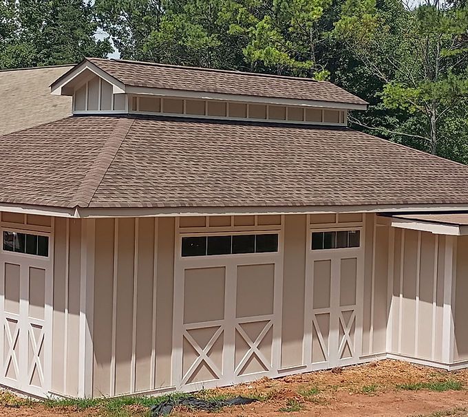 White barn with tan trim and brown shingled roof in a wooded yard
