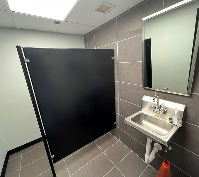 Public restroom stall with black partition, sink, mirror, and gray tiled walls and floor